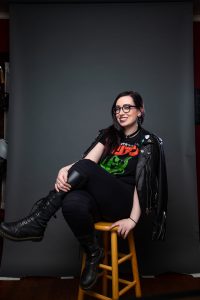SWRolodex new owner Merrick Monroe smiles while sitting on a wooden stool, legs crossed, wearing classes, a leather jacket with pins, and black clothing.photo by Joshua Dommermuth of Quixotic Images
